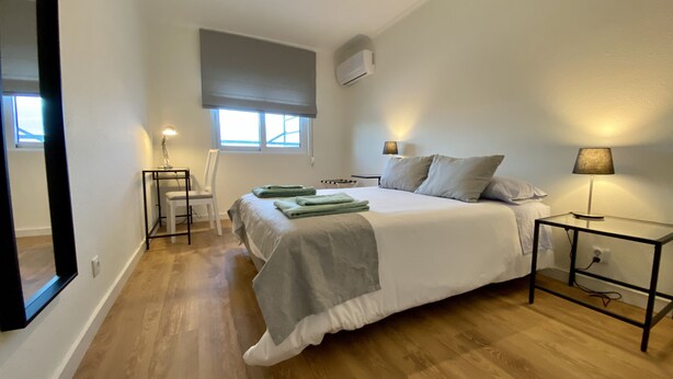 A bedroom with a bed, bedside tables, a chair, a window, and an air conditioning unit.