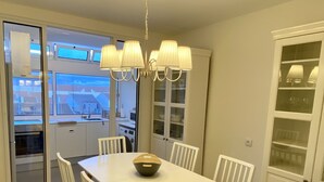 A dining area with a white table and chairs, a hanging light fixture, and a view of the outside through a glass door.