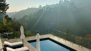 A modern outdoor pool area with lounge chairs and umbrellas, surrounded by a glass railing, overlooking a scenic hillside. - West Hollywood Hills Pool Home With Views (Los Angeles)