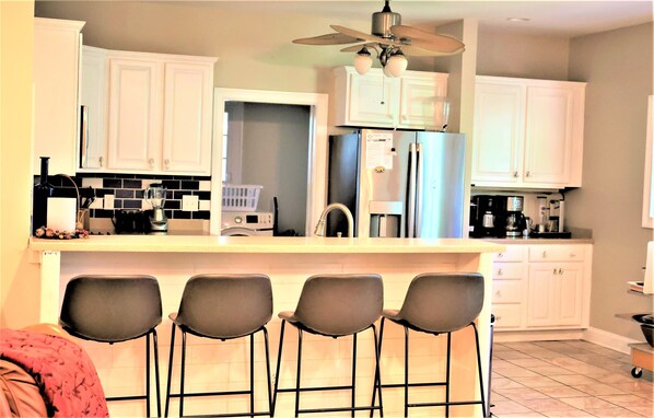 A kitchen with white cabinets, a black countertop, and a ceiling fan. There are four bar stools in front of the counter.