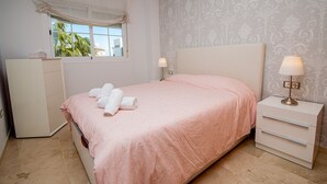Quarto com cama, mesa de cabeceira, abajur, guarda-roupa e janela com vista para palmeiras.