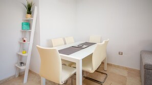A white dining table with four chairs, a plant on a shelf, and a book titled 'Grown Ups' on the shelf.
