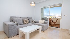 Uma sala de estar moderna com um sofá cinzento, almofadas estampadas, uma mesa de centro branca e vista para o exterior através de uma porta de vidro.