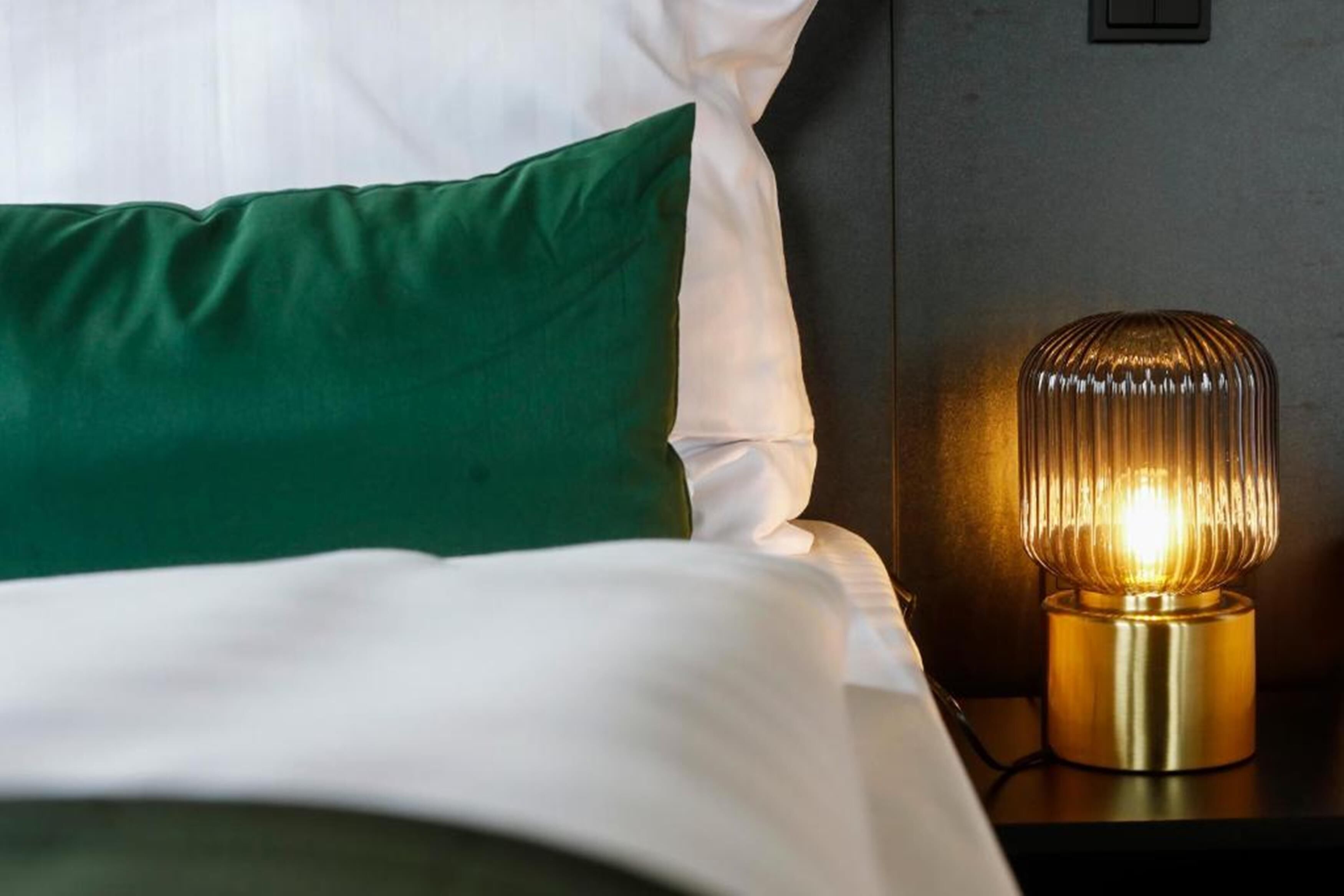 A neatly made bed with white and green bedding, a modern lamp on a bedside table, and a dark wall in the background.