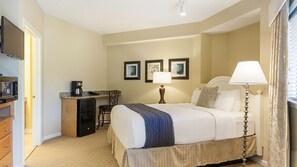 Ein Hotelzimmer mit einem Bett, Nachttischlampen, einem Schreibtisch mit Kaffeemaschine, einem Fernseher und gerahmten Bildern an der Wand.