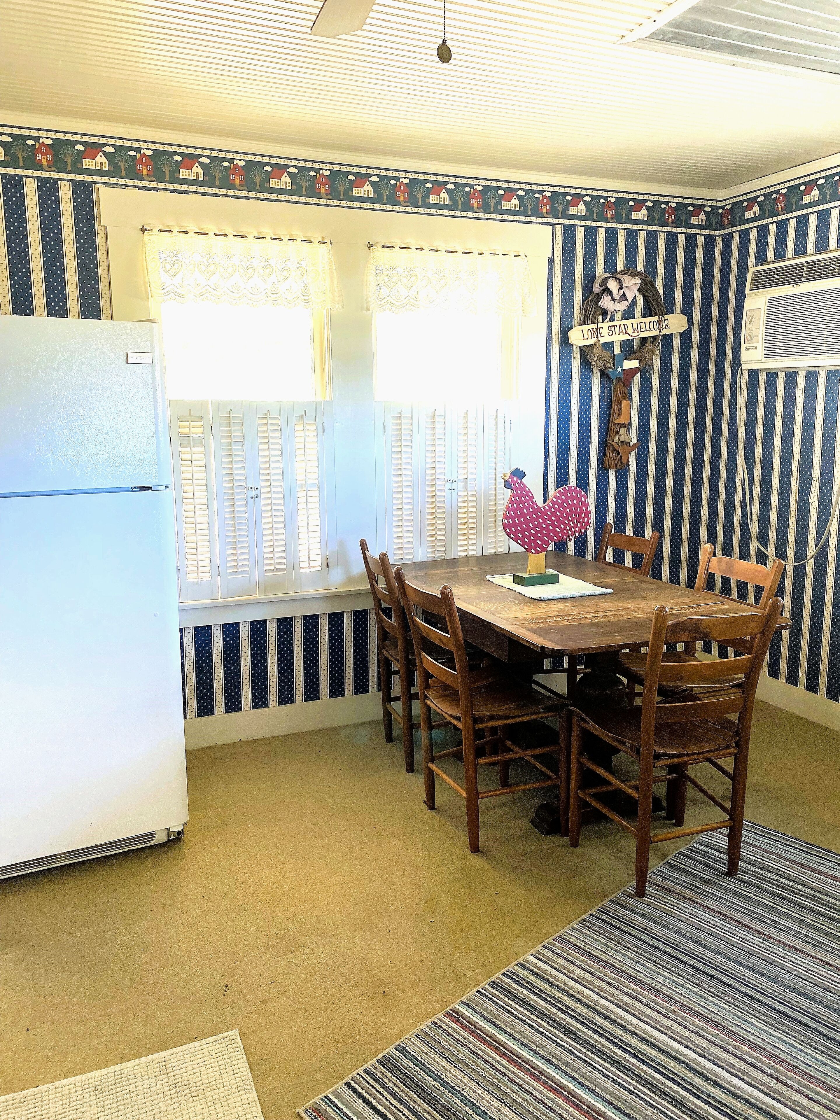 A kitchen with a dining table, chairs, a refrigerator, and a window with curtains.