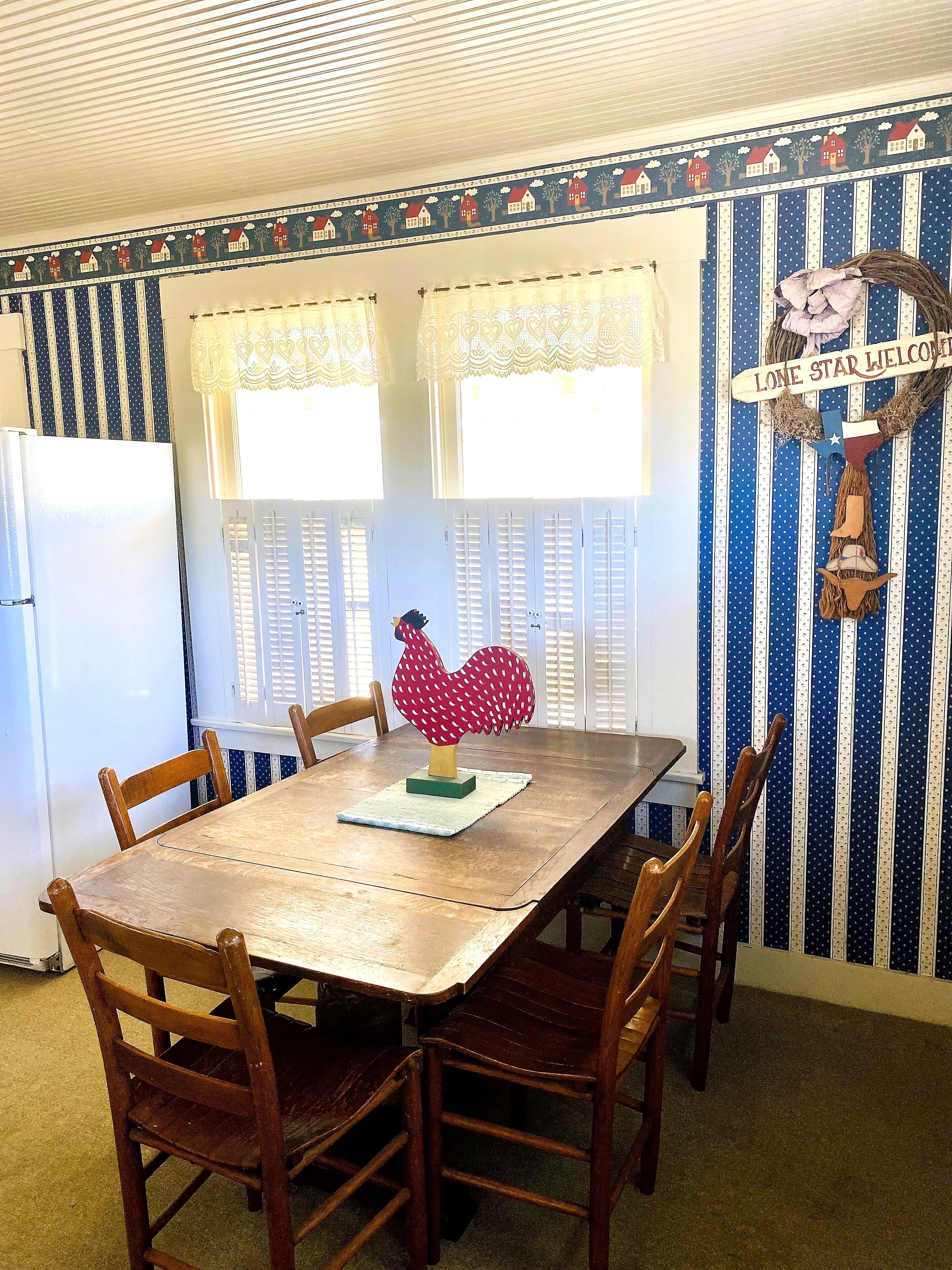 A dining area with a wooden table and chairs, a decorative rooster centerpiece, and striped wallpaper with a 'Lone Star Welcome' sign.