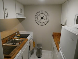 A compact kitchen with white cabinets, a stainless steel sink, a stove, and a trash bin.