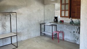 Eine Küche mit Betonboden, einem Metallregal, einem roten Hocker, einem Holzkasten mit Glastür und einer Pendelleuchte.