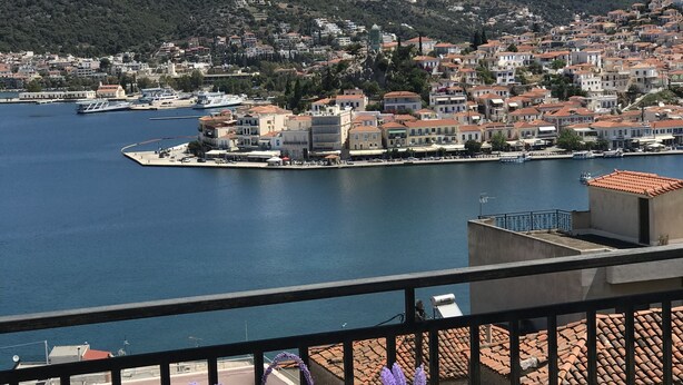 Un balcon avec une table dressée pour deux personnes, offrant une vue sur une ville côtière, avec une vue dégagée sur la mer et les collines en arrière-plan.