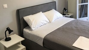 Un lit bien fait, avec une literie blanche et grise, une lampe de chevet, une petite table de chevet blanche avec un tiroir, un tableau encadré au mur et un appareil de climatisation au-dessus du lit.