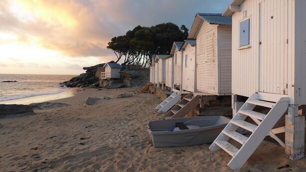 Cabànes de fusta a peu de platja amb escales exteriors, un vaixell i una riba rocosa.