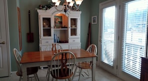 Une salle à manger avec une table en bois et quatre chaises, un buffet blanc aux portes vitrées et une fenêtre avec des stores.