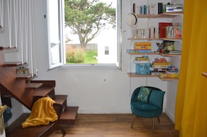 A room with a wooden staircase, a window with a view of a tree, a teal chair, and a bookshelf with various books and boxes.