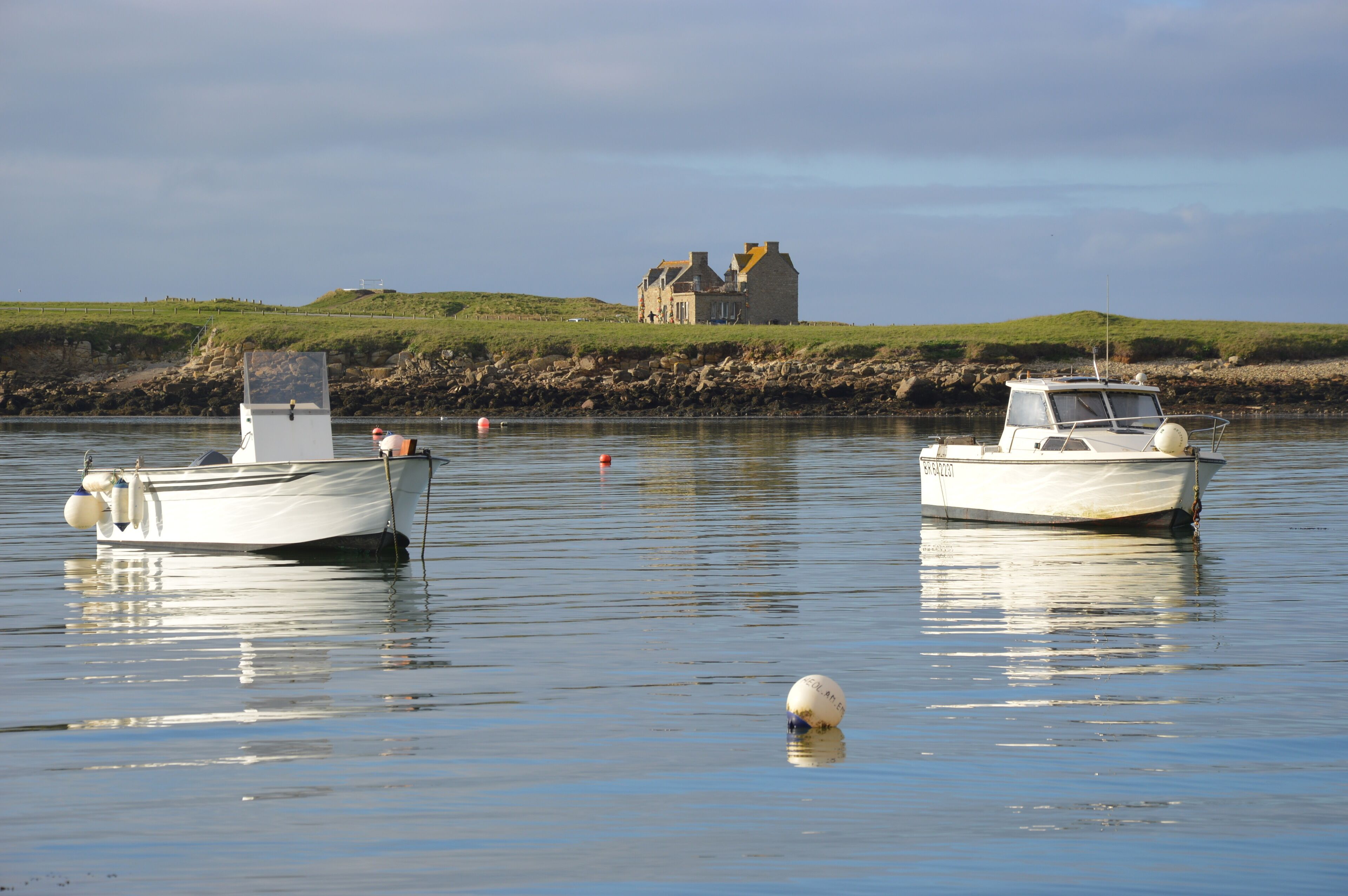 Two boats anchored near a coastal house with a grassy hill in the background.