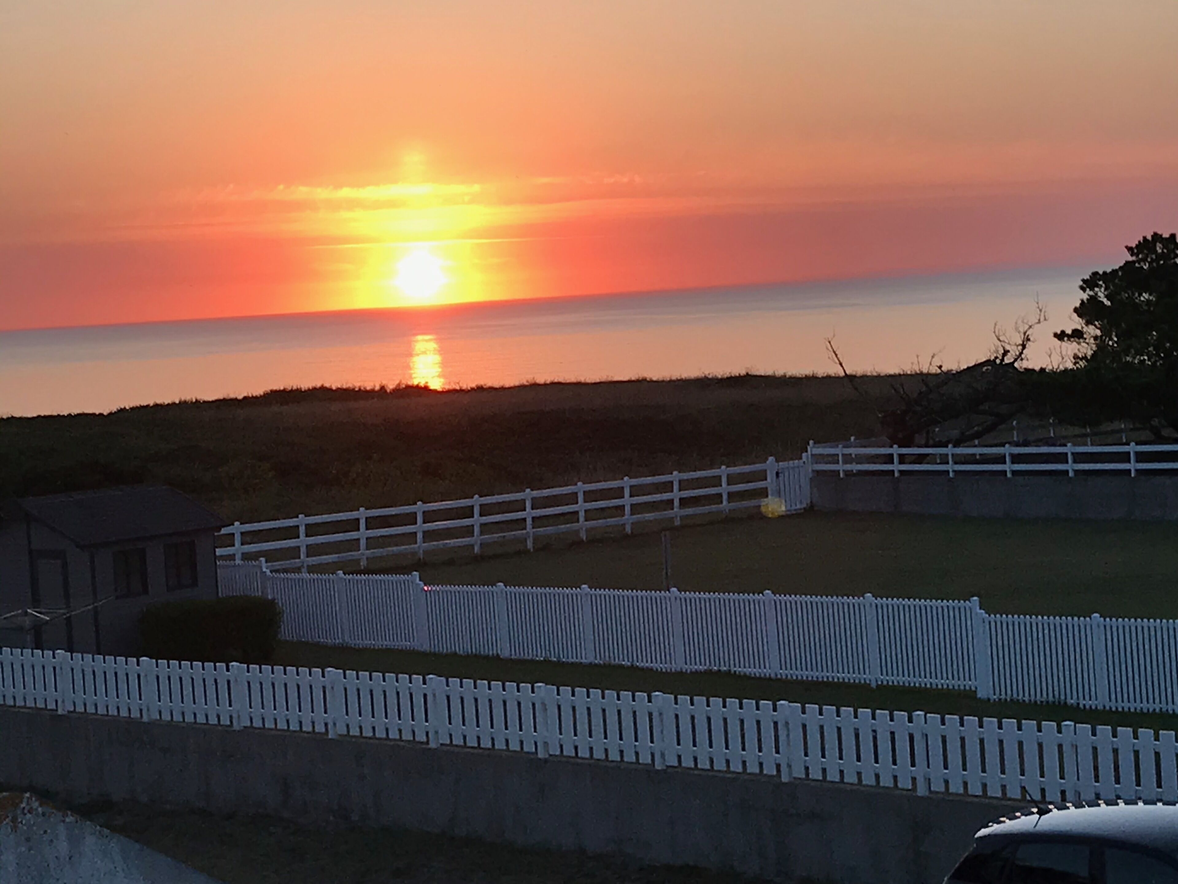 A sunset over a coastal area with a white fence and a building in the foreground.