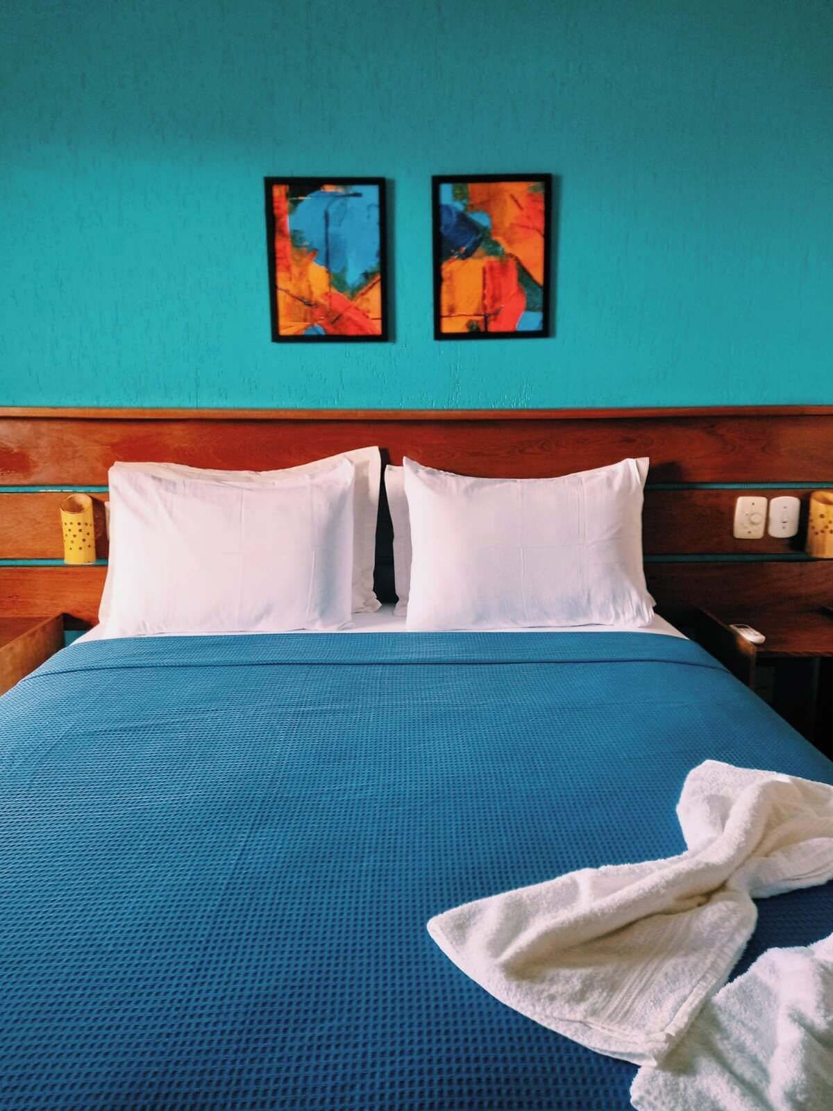 Una cama con cubrecama azul, dos almohadas blancas y un albornoz blanco en el suelo. Encima de la cama, hay dos cuadros abstractos en la pared.