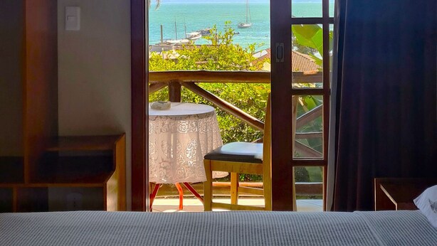 A hotel room with a bed, a ceiling fan, a window with a view of the sea and mountains, and an air conditioning unit. - Pousada Aquarela do Mar (Angra dos Reis)