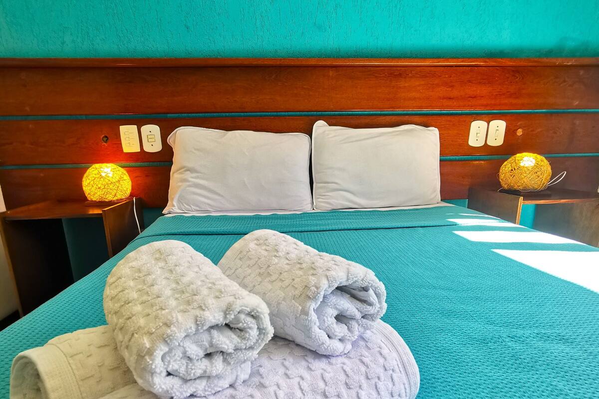 A neatly made bed with white pillows and rolled towels, a wooden headboard, and a teal wall.