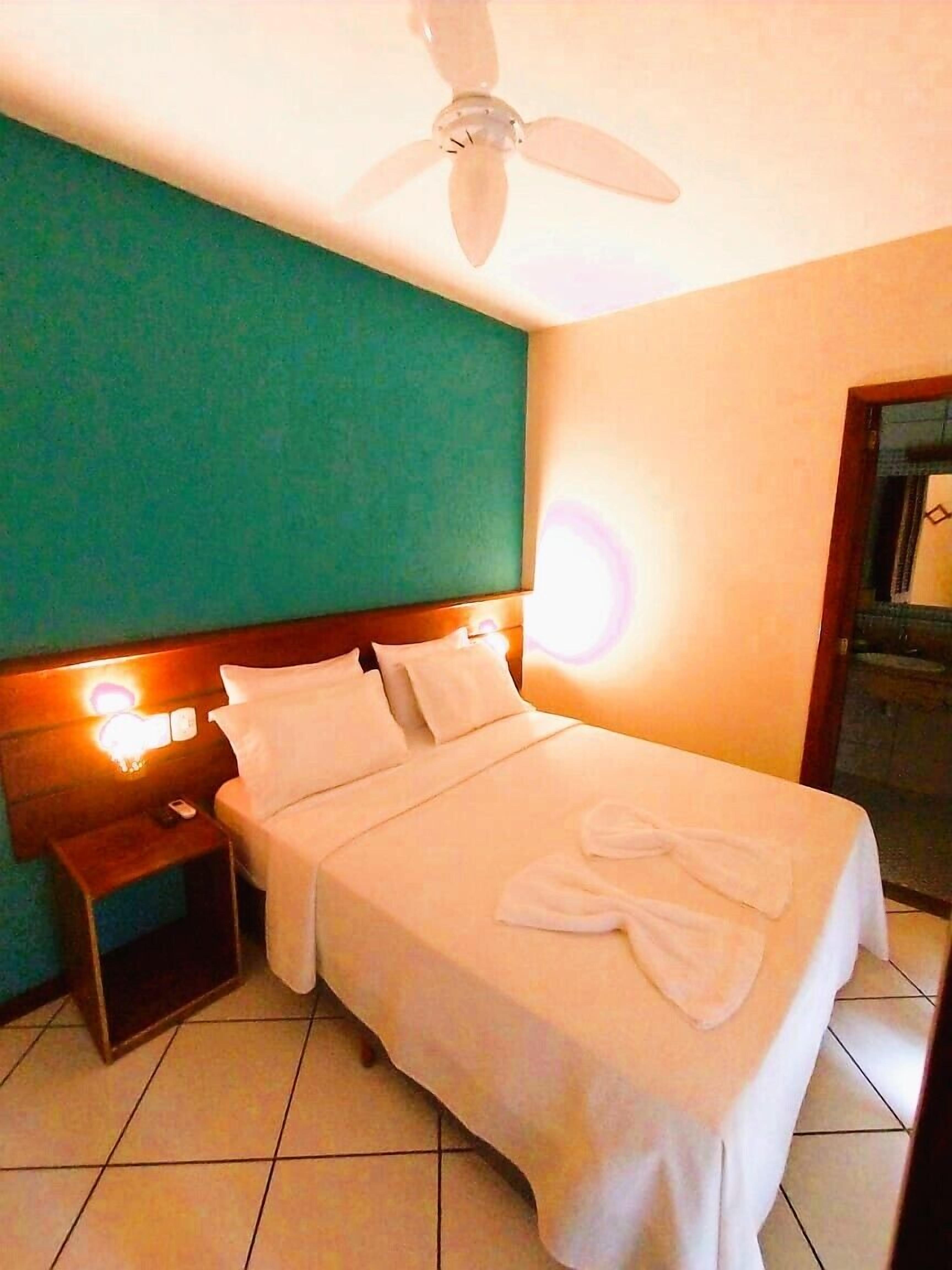 Habitación de hotel con cama, mesita de noche, ventilador de techo y lámpara en la pared.