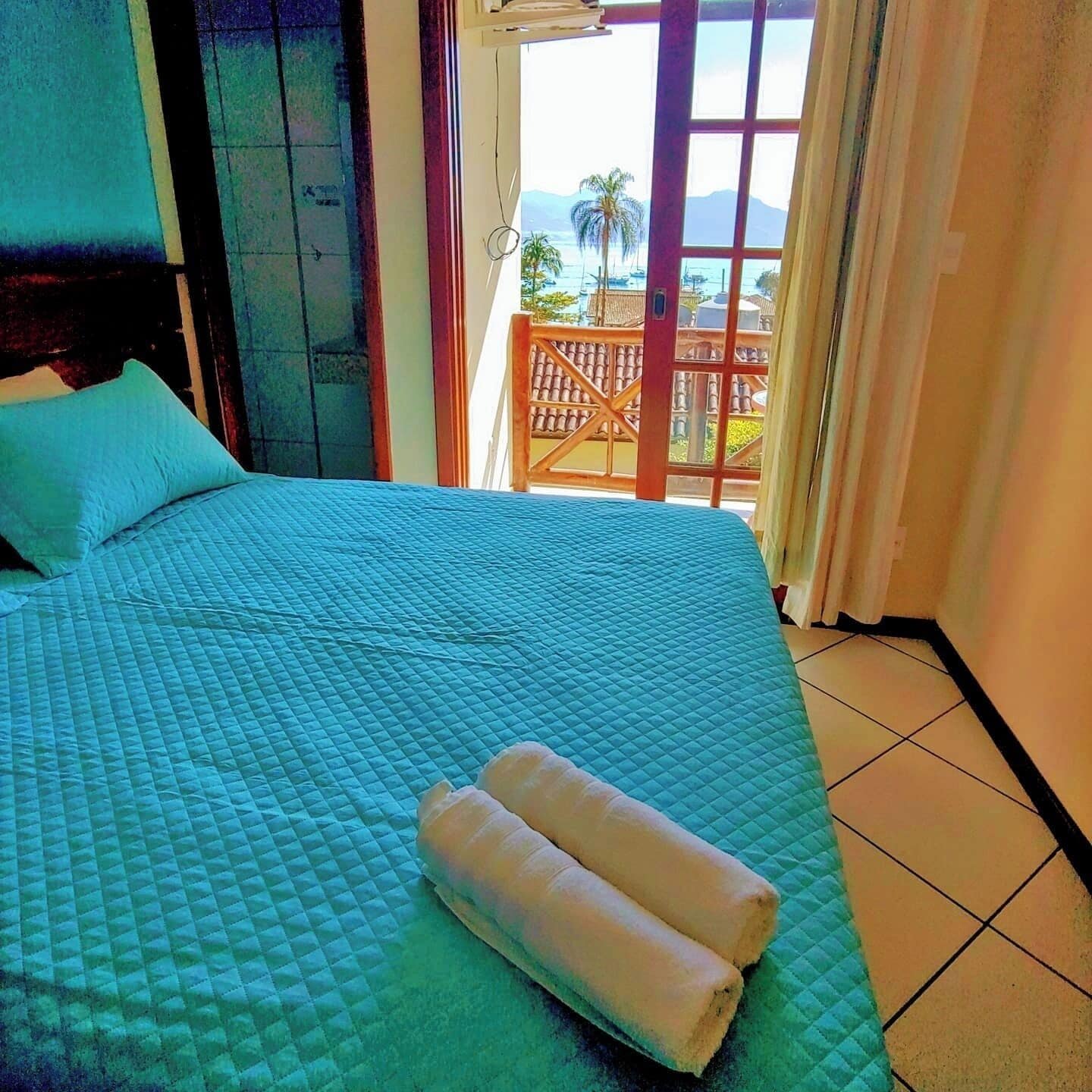 A bed with a quilted blue bedspread, two rolled towels, and a view of a balcony with a railing and palm trees.
