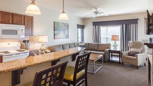 A modern kitchen with a built-in island, a dining area with a table and chairs, a seating area with a sofa and armchairs, and a large window with blinds. - One Bedroom, Luxury Condo, Williamsburg Parkside, Virginia (2629717) (Williamsburg)