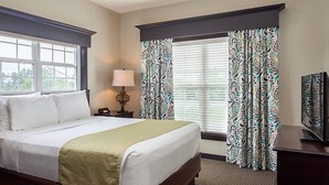 A hotel room with a bed, a nightstand with a lamp, a television, and patterned curtains. - One Bedroom, Luxury Condo, Williamsburg Parkside, Virginia (2629717) (Williamsburg)