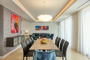 Une salle à manger moderne avec une longue table, des chaises noires et un lustre. Les murs sont ornés de peintures abstraites et une grande fenêtre est agrémentée de rideaux.