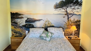 Un dormitorio con cama, dos mesitas de noche, una lámpara y un mural con paisaje costero.