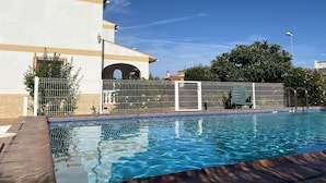 Una piscina con borde de azulejos, un edificio blanco con una franja color beige y una cerca de metal.