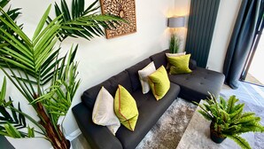 Una sala moderna con un sofá modular, cojines decorativos, una obra de arte en la pared, una lámpara de pie y plantas verdes grandes.