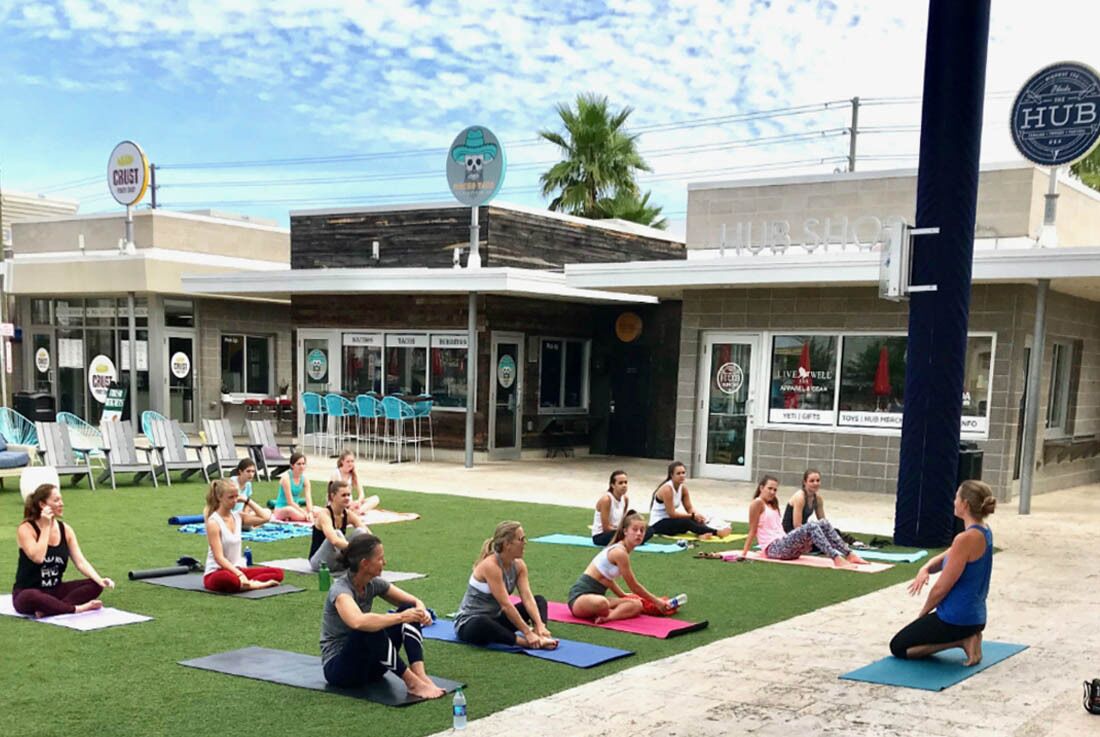 Un groupe de personnes, sur des tapis de yoga, pratiquent en plein air devant un bâtiment sur lequel on peut lire l’inscription « VIVEZ MIEUX ».