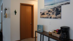 A hallway with a wooden door, a guitar, a framed beach picture, a small table with a coffee maker, and a basket of snacks. - Canal front Island Escape for the family! Dog-Friendly with private Dock  (Ocean Shores)