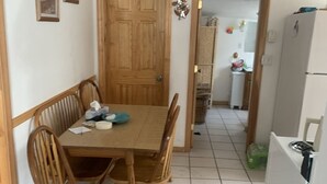 Zona pranzo con tavolo e sedie in legno, una ventola a soffitto e una cucina visibile attraverso un'apertura.