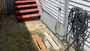 Un piccolo edificio a un piano con una porta rossa e una finestra bianca, una scala in legno e un tubo da giardino avvolto contro il muro.