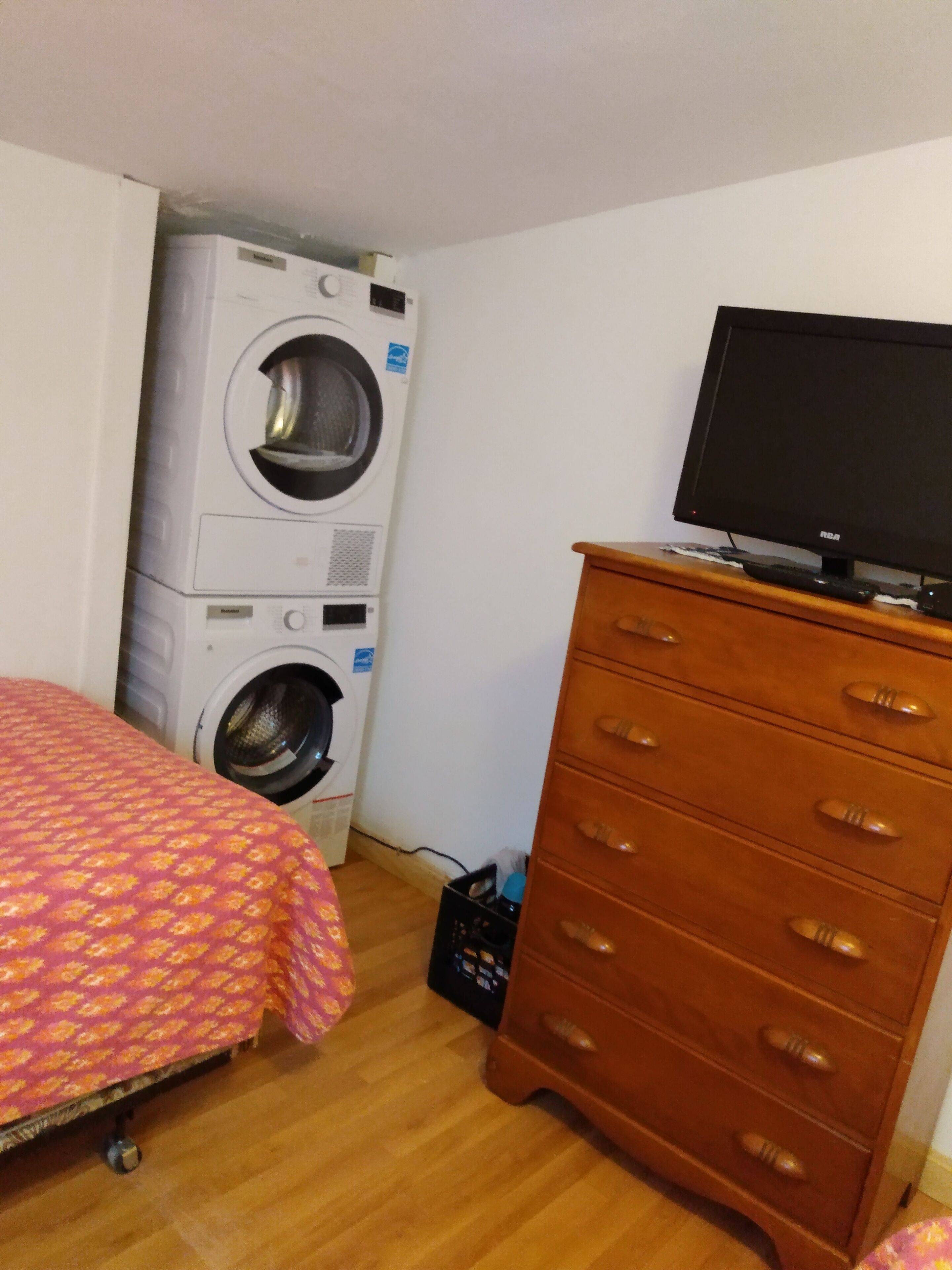 Een slaapkamer met een wasmachine, een televisie op een dressoir en een bed met een gestreept beddegoed.