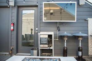 A building with a grey exterior, a fire extinguisher mounted on the wall, a television mounted above a small white cabinet, and two outdoor heaters. - Cheerful Townhome Sloan’s Lake (Denver)