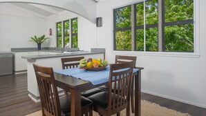 A modern kitchen with a dining area, large windows, and a fruit bowl on the table.