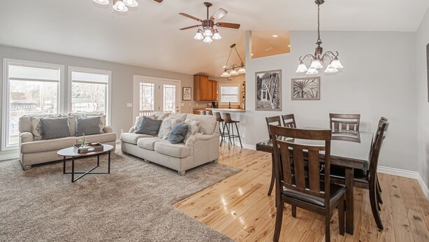 A spacious living room with a large window, a dining area with wooden chairs, and a kitchen in the background.