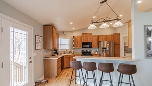 A modern kitchen with wooden cabinets, a stainless steel refrigerator, a large island with bar stools, and pendant lighting.