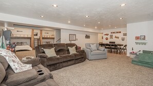 A spacious living area with a brown sofa, a grey armchair, and a green sofa. There is a dining table with chairs and a bunk bed in the background.