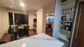 Una cocina moderna con encimera de mármol, una zona de comedor con mesa oscura y un televisor al fondo.