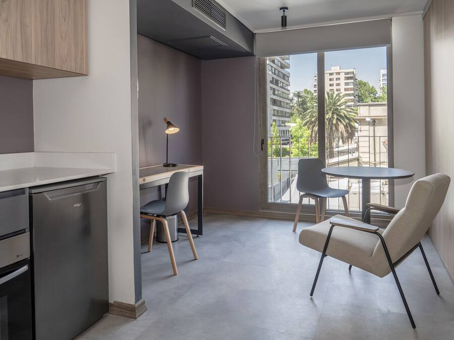 A modern kitchen with a built-in dishwasher, a dining area with a round table and two chairs, and a large window offering a view of the cityscape.