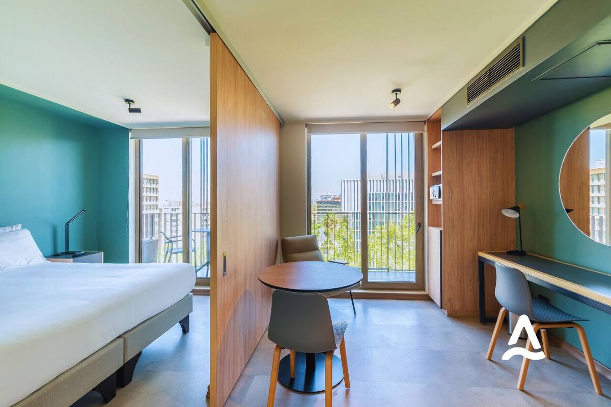 Een moderne hotelkamer met een bed, een ronde tafel met stoelen, een bureau en een groot raam met uitzicht op de stad.