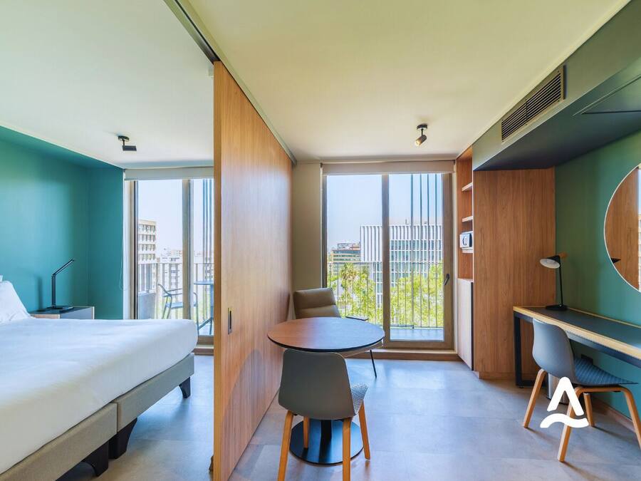 Una habitación de hotel moderna con una cama, una mesa redonda con sillas, un escritorio y un amplio ventanal con vista a la ciudad.