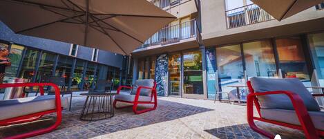 A modern outdoor seating area with red-framed chairs and a patterned floor, overlooking a building with large windows and a balcony.
