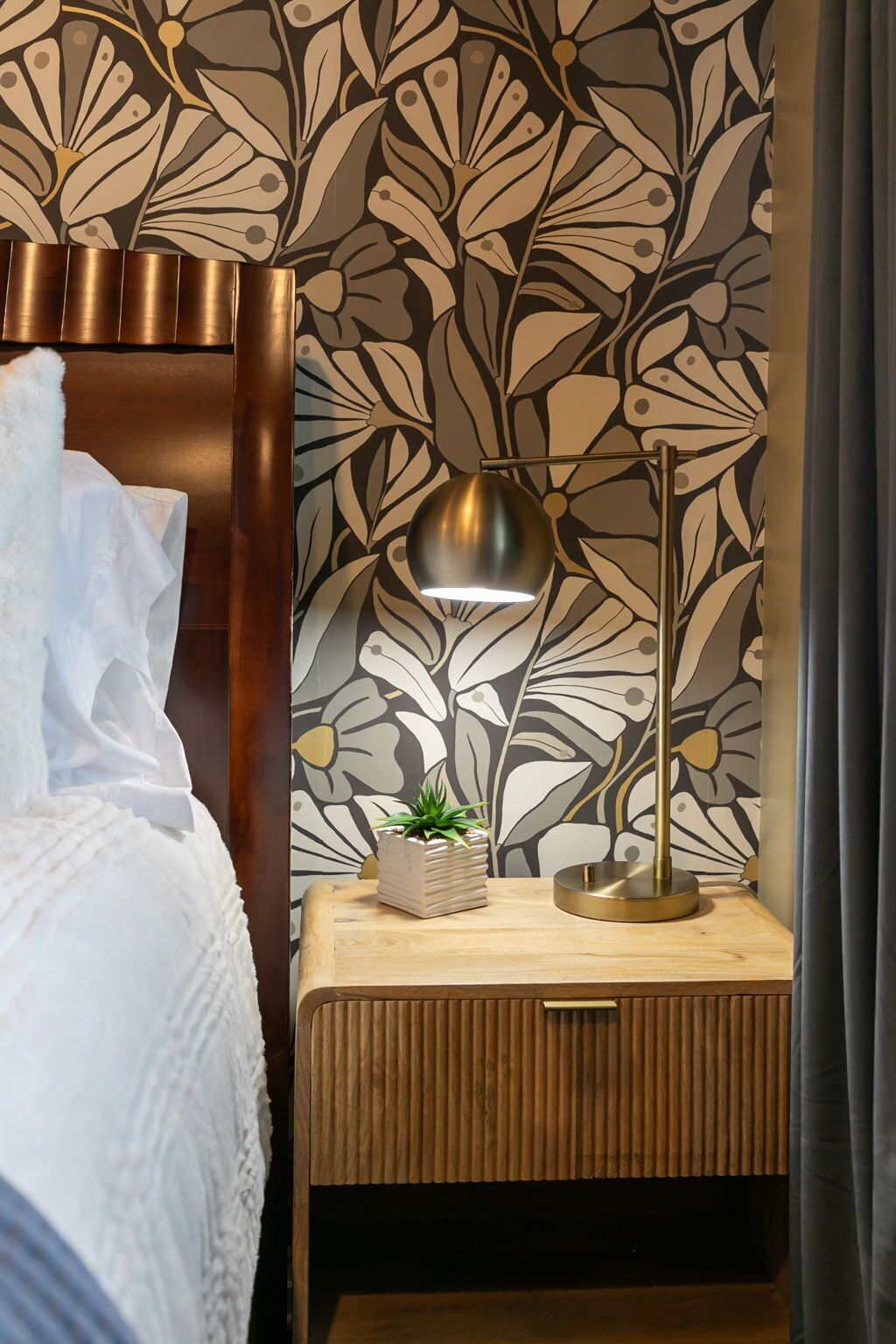 A neatly made bed with a white duvet, a bedside table with a small potted plant, a modern floor lamp, and a wall with a floral pattern.