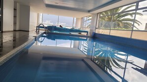 An indoor swimming pool with clear blue water, large windows, and a view of palm trees outside. - 500 Excellent panoramic view, olympic stadium, park, mountain swimming pool (Quito)