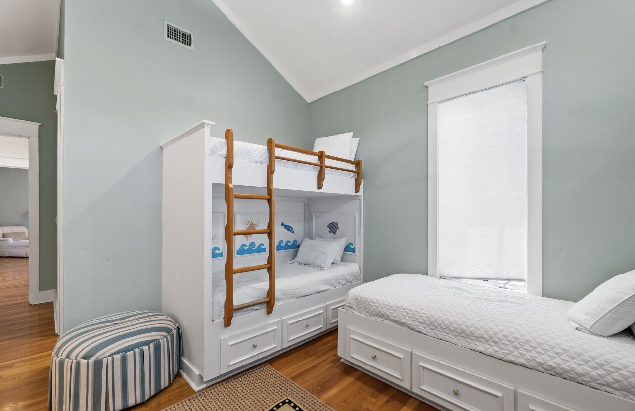 A bedroom with a bunk bed, a single bed, a window with blinds, a striped ottoman, and a wooden ladder.