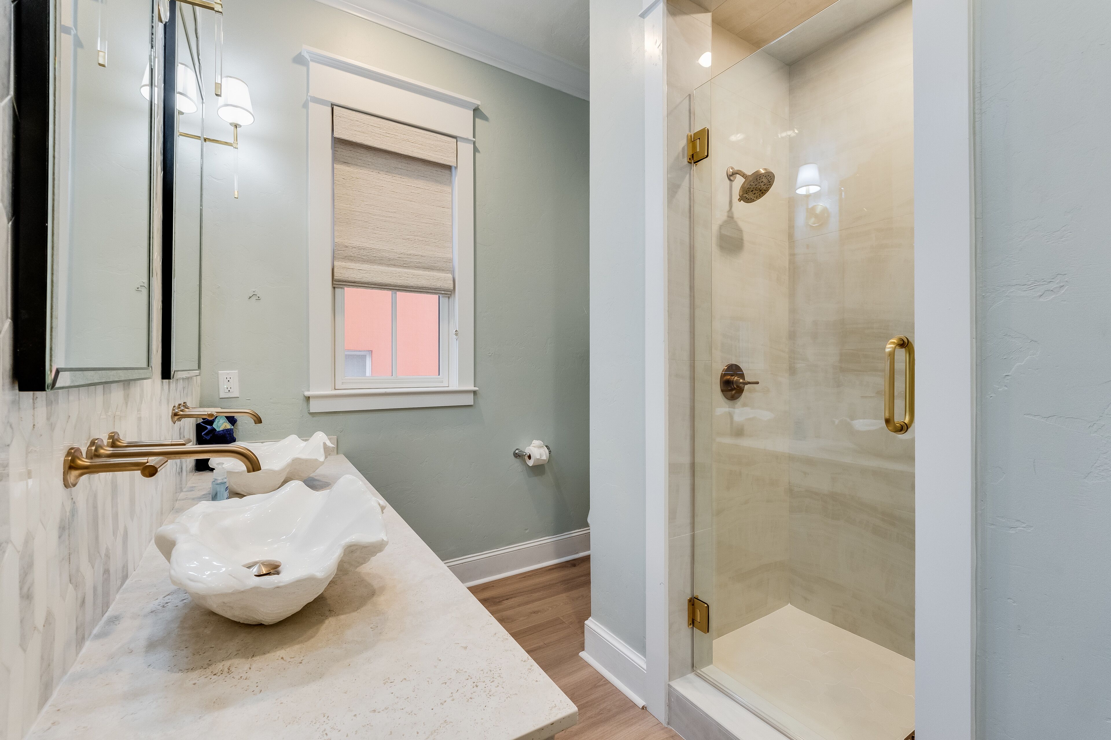 A bathroom with a marble vanity, a large white bowl sink, a glass shower enclosure, and a window with a beige blind.